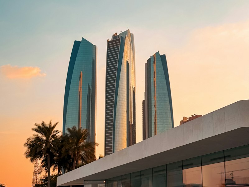 Three curved glass towers reflecting an orange sunset sky above palm trees and a low modern building in Abu Dhabi