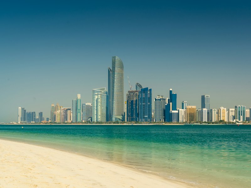 Abu Dhabi skyline of glass towers seen across turquoise water from a sandy beach under a clear blue sky