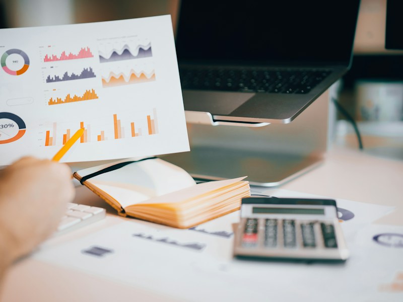 Person reviewing printed financial charts beside a laptop and calculator, representing CRS and CARF reporting preparation