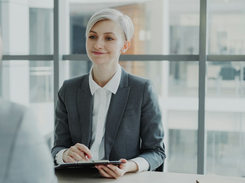 Interviewer in a grey suit holding a clipboard across a desk from a job candidate in a bright office