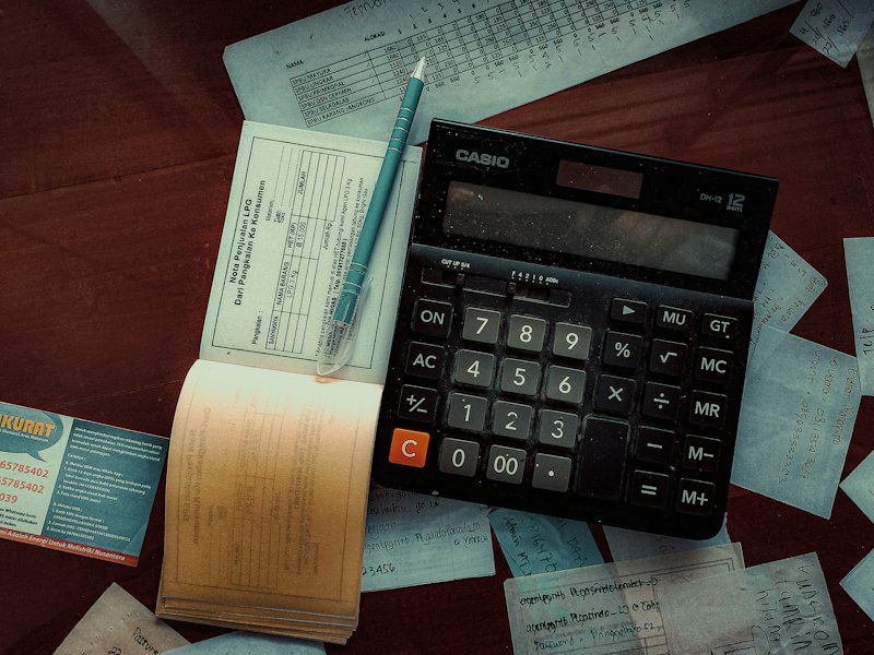 Black desktop calculator and a teal pen resting on an open receipt book, with handwritten notes and paper slips spread across a dark wooden desk
