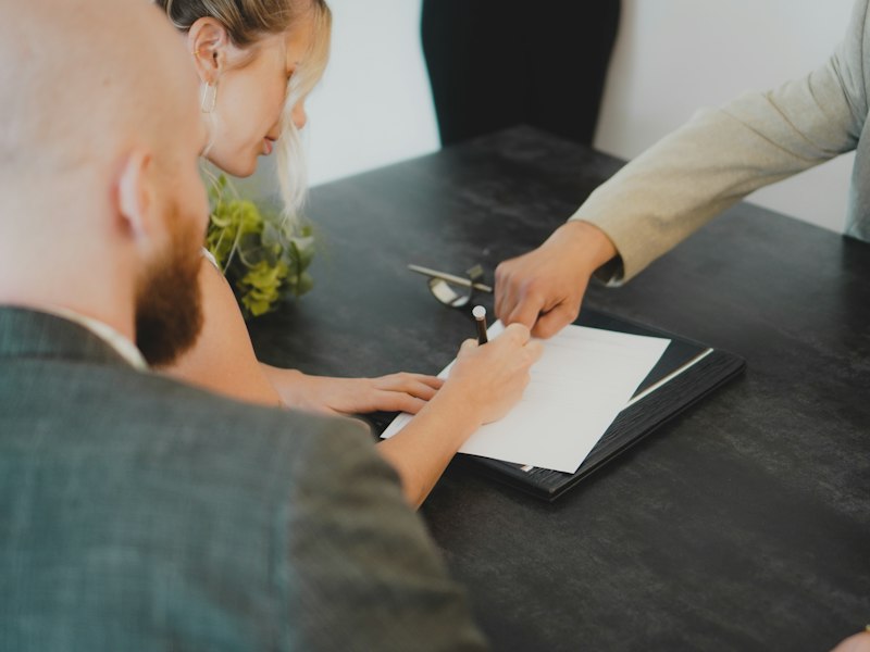 A woman signs a document at a dark desk while a man beside her and another person across the table look on and point to the page