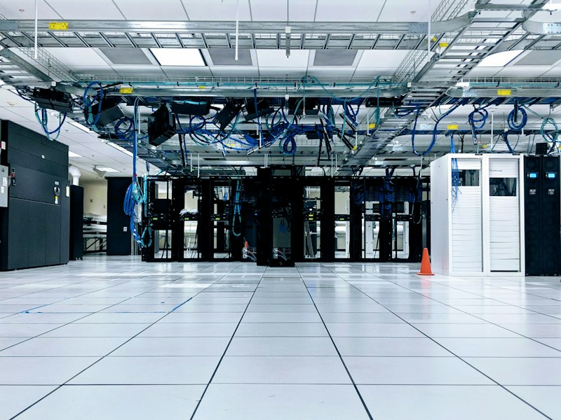 Data centre hall with rows of black server racks, white cabinets and blue network cables running along overhead trays