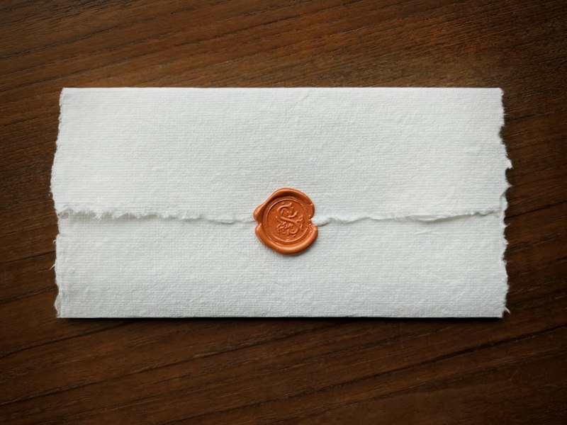 Folded white handmade paper envelope closed with an orange wax seal, on a dark wooden table