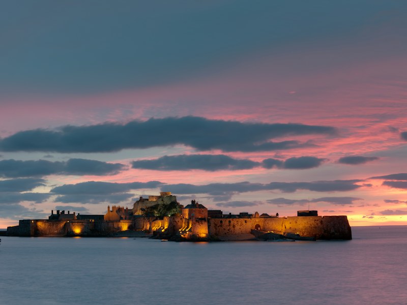 A floodlit island fortress on calm water under a pink dusk sky