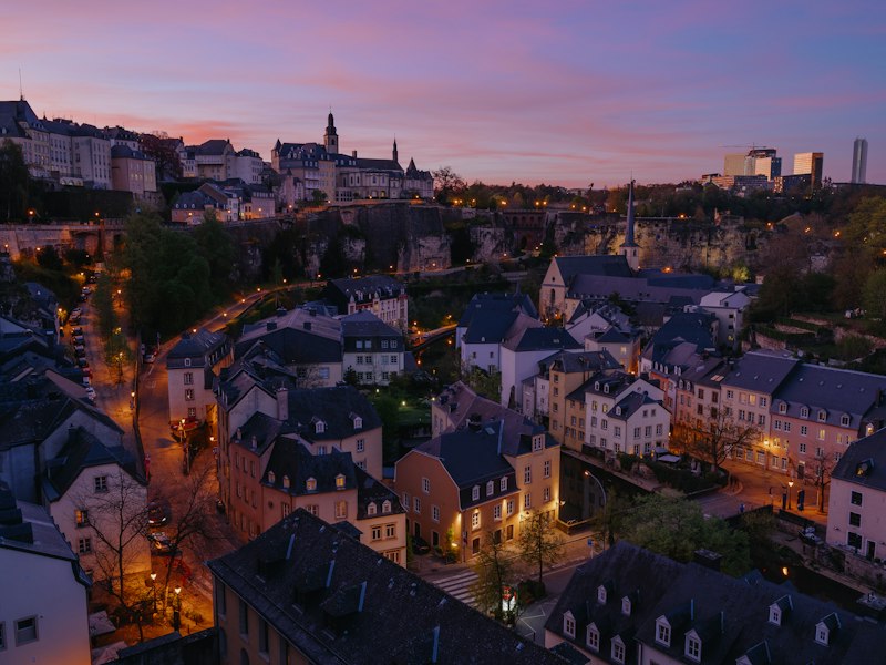 Grund district of Luxembourg City at dusk, with lit old houses in the valley below the city walls and Kirchberg office towers on the skyline