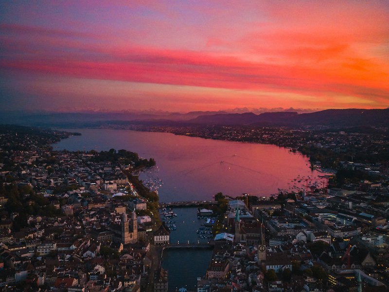 Aerial view of Zurich at sunset, with the Limmat river flowing from Lake Zurich between old town buildings and church towers, and hills on the horizon