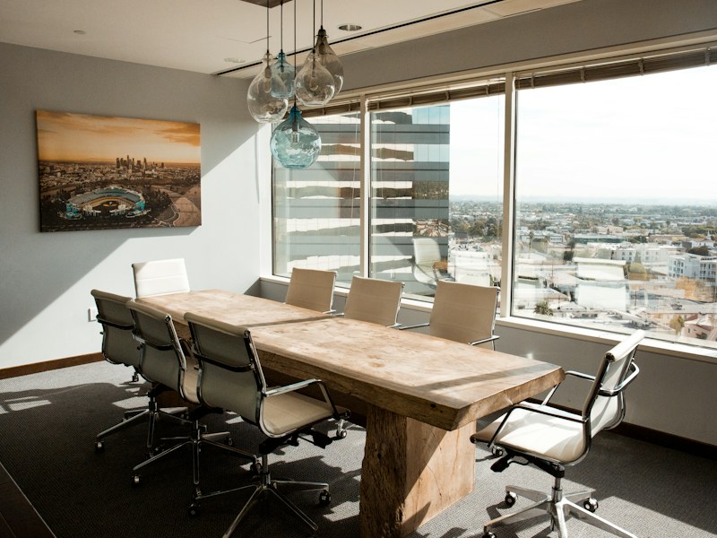 Empty boardroom with a wooden conference table and white chairs beside large city-view windows