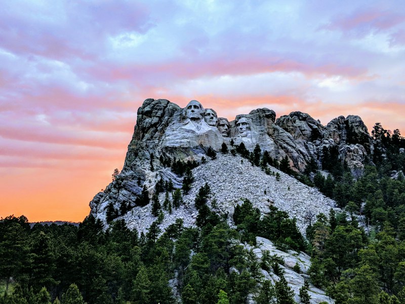 Mount Rushmore's carved granite faces above a pine-covered slope under a pink and orange sunset sky in South Dakota