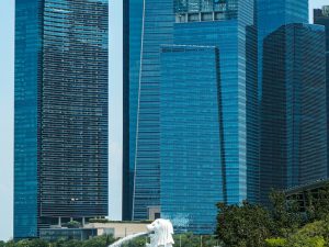Merlion statue spouting water in front of blue glass office towers in Singapore's Marina Bay financial district